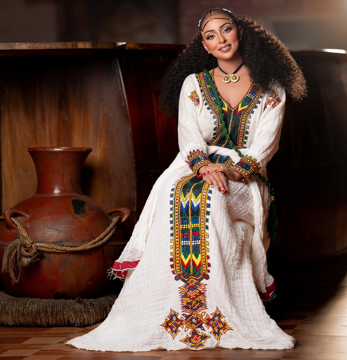 A woman wearing a traditional white embroidered long Ethiopian National Dress with colorful geometric patterns sits in a warm indoor setting next to a large terracotta pot.