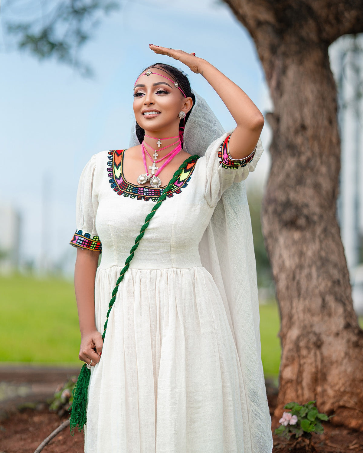 Woman in traditional Axum Fetil with a green scarf, standing outdoors with trees and grass in the background.