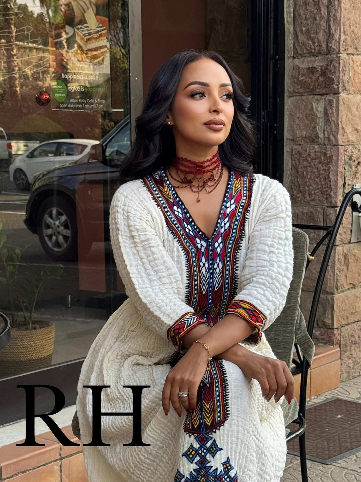 A woman wearing a white textured Axum Fetil dress with colorful geometric embroidery, sitting outdoors in front of a storefront. The image features a large black 'RH' watermark overlay in the bottom left corner.