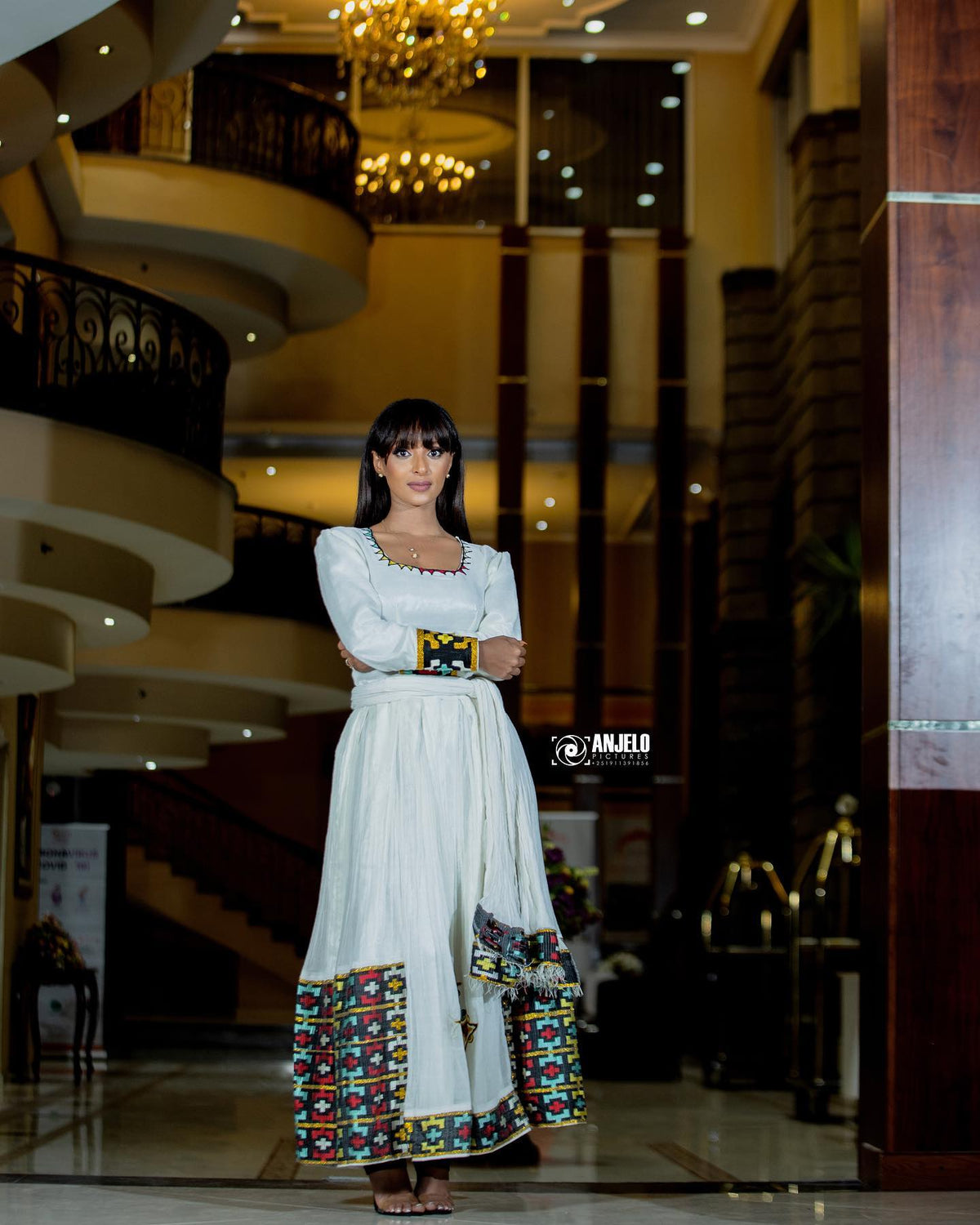 A woman wearing a long white traditional Axum Fetil with colorful embroidered patterns at the hem and cuffs, standing in a grand hotel lobby with chandeliers and curved balconies in the background.