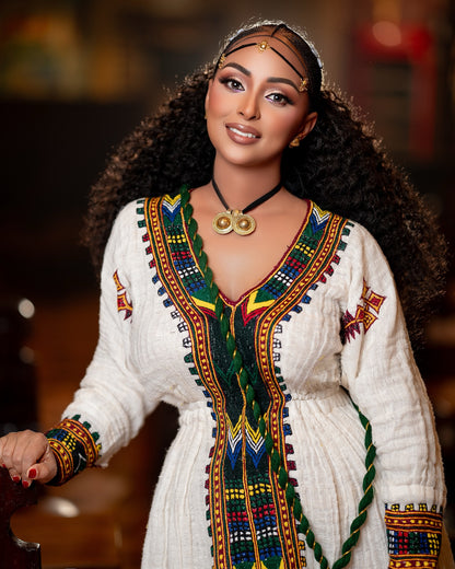 A woman wearing a white embroidered traditional Ethiopian National Dress with colorful geometric patterns, a black necklace with gold medallions, and a headpiece, posed against a warm, blurred background.
