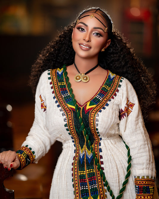 A woman wearing a white embroidered traditional Ethiopian National Dress with colorful geometric patterns, a black necklace with gold medallions, and a headpiece, posed against a warm, blurred background.