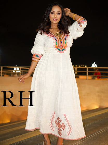 A woman modeling a white midi Ethiopian National Dress with colorful embroidered details on the bodice and sleeves, set against a nighttime outdoor background with a large 'RH' watermark on the left.