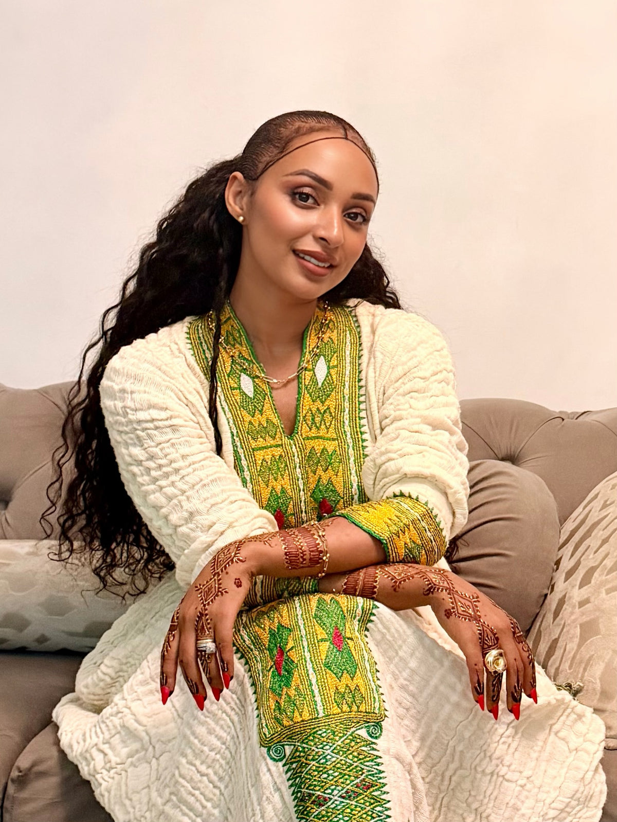 A woman with long, curly dark hair sits on a beige sofa wearing a cream-colored textured cardigan over a vibrant green and yellow embroidered traditional Axum Fetil. She has intricate henna designs on her hands and arms, and is wearing gold jewelry.