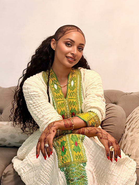 A woman with long, curly dark hair sits on a beige sofa wearing a cream-colored textured cardigan over a vibrant green and yellow embroidered traditional Axum Fetil. She has intricate henna designs on her hands and arms, and is wearing gold jewelry.
