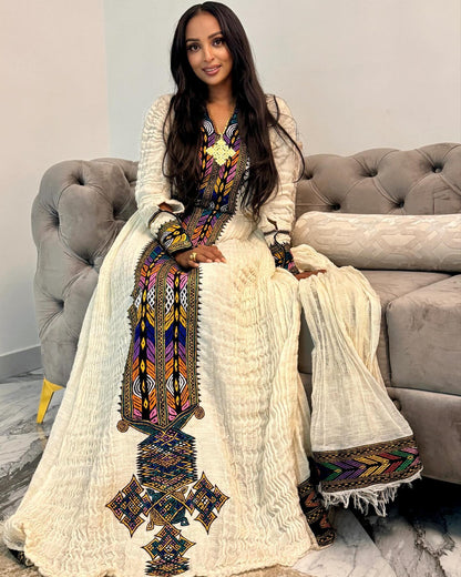 A woman wearing a long cream-colored Moroccan Ethiopian National Dress with colorful geometric embroidery, sitting on a gray tufted sofa.