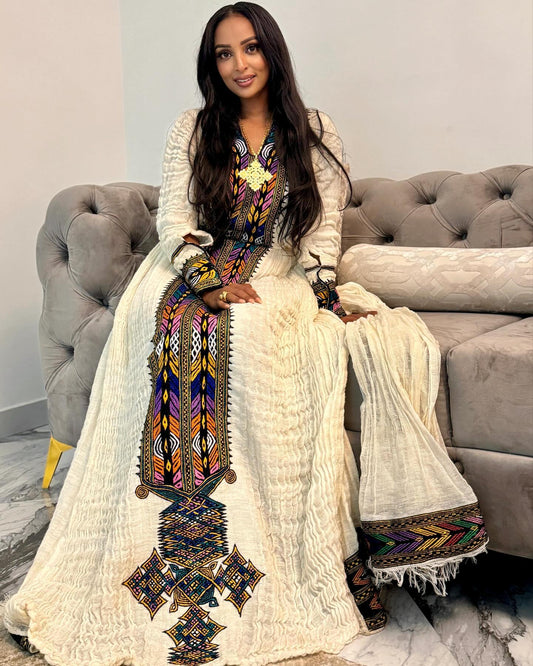 A woman wearing a long cream-colored Moroccan Ethiopian National Dress with colorful geometric embroidery, sitting on a gray tufted sofa.