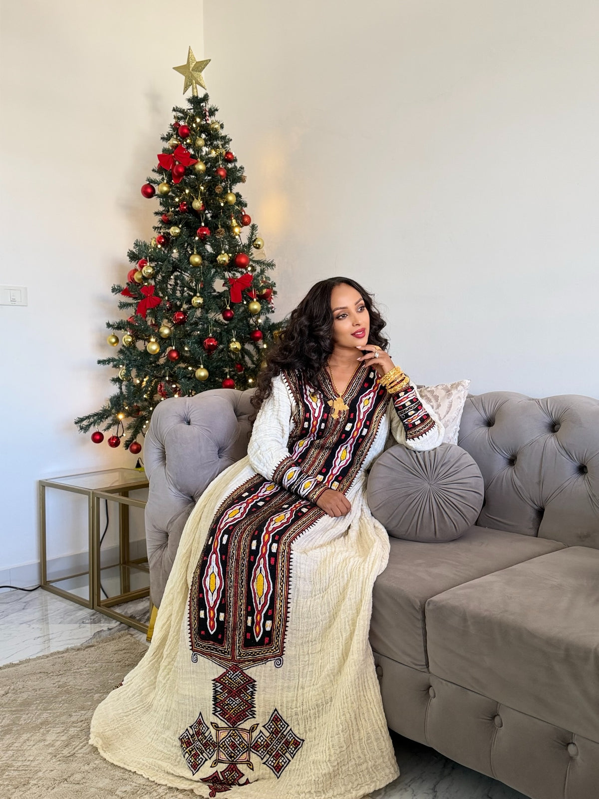 A woman wearing a traditional long white Axum Fetil with a colorful embroidered vest sits on a gray tufted sofa next to a decorated Christmas tree.