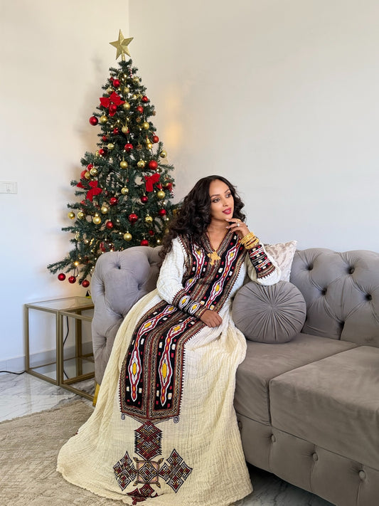 A woman wearing a traditional long white Axum Fetil with a colorful embroidered vest sits on a gray tufted sofa next to a decorated Christmas tree.