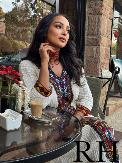 A woman seated at an outdoor cafe table wearing a white textured cardigan over a colorful geometric print Axum Fetil. She is holding a glass of coffee, with a stone wall, glass windows, and a potted red flower in the background.