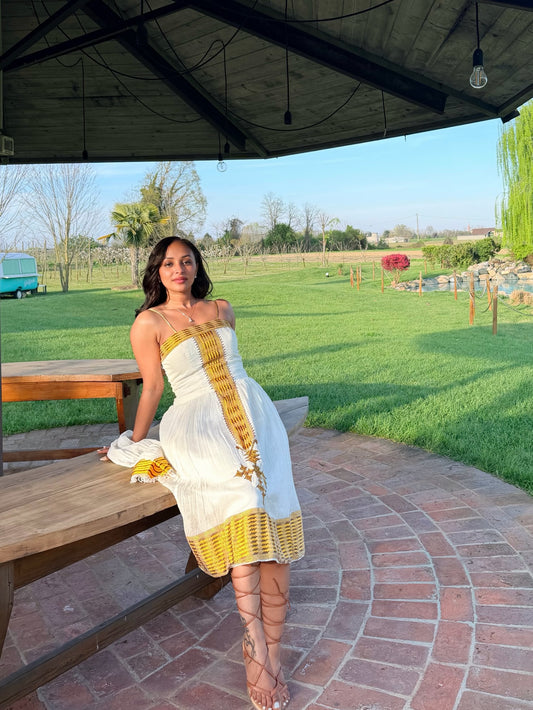 A woman wearing a white strapless Ethiopian National Dress with gold embroidered details and lace-up heels, posing on a brick patio under a wooden pavilion next to a wooden bench in a grassy outdoor setting.