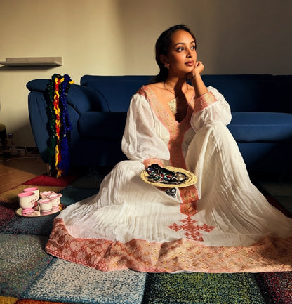 A woman sits on a colorful rug in front of a blue sofa, wearing a white embroidered Ethiopian National Dress with pink and peach detailing. She holds a tray with dark sunglasses, and small pink cups sit on a side table to her left.