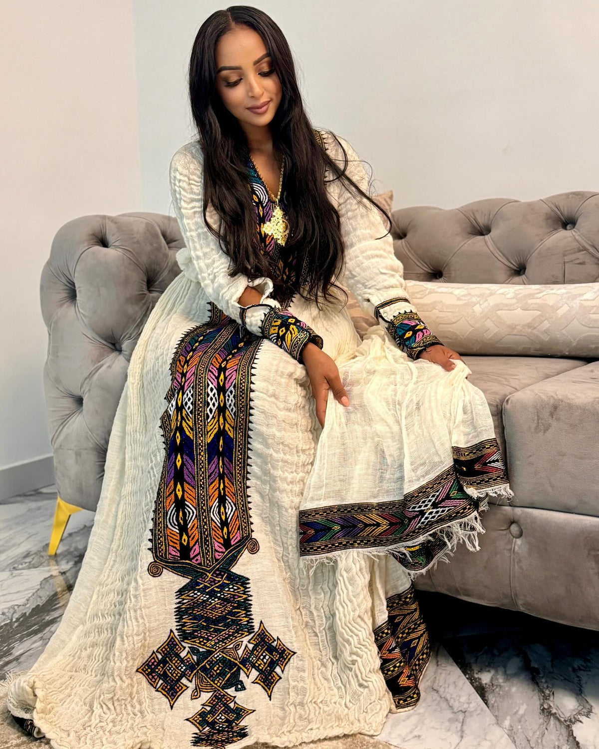 A woman wearing a long white embroidered Ethiopian National Dress with colorful geometric patterns, seated on a gray tufted sofa in an indoor setting.