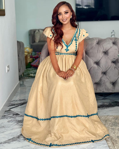 A woman wearing a beige formal Axum Fetil with turquoise blue embroidery and beadwork, seated in a living room with a gray tufted sofa in the background.