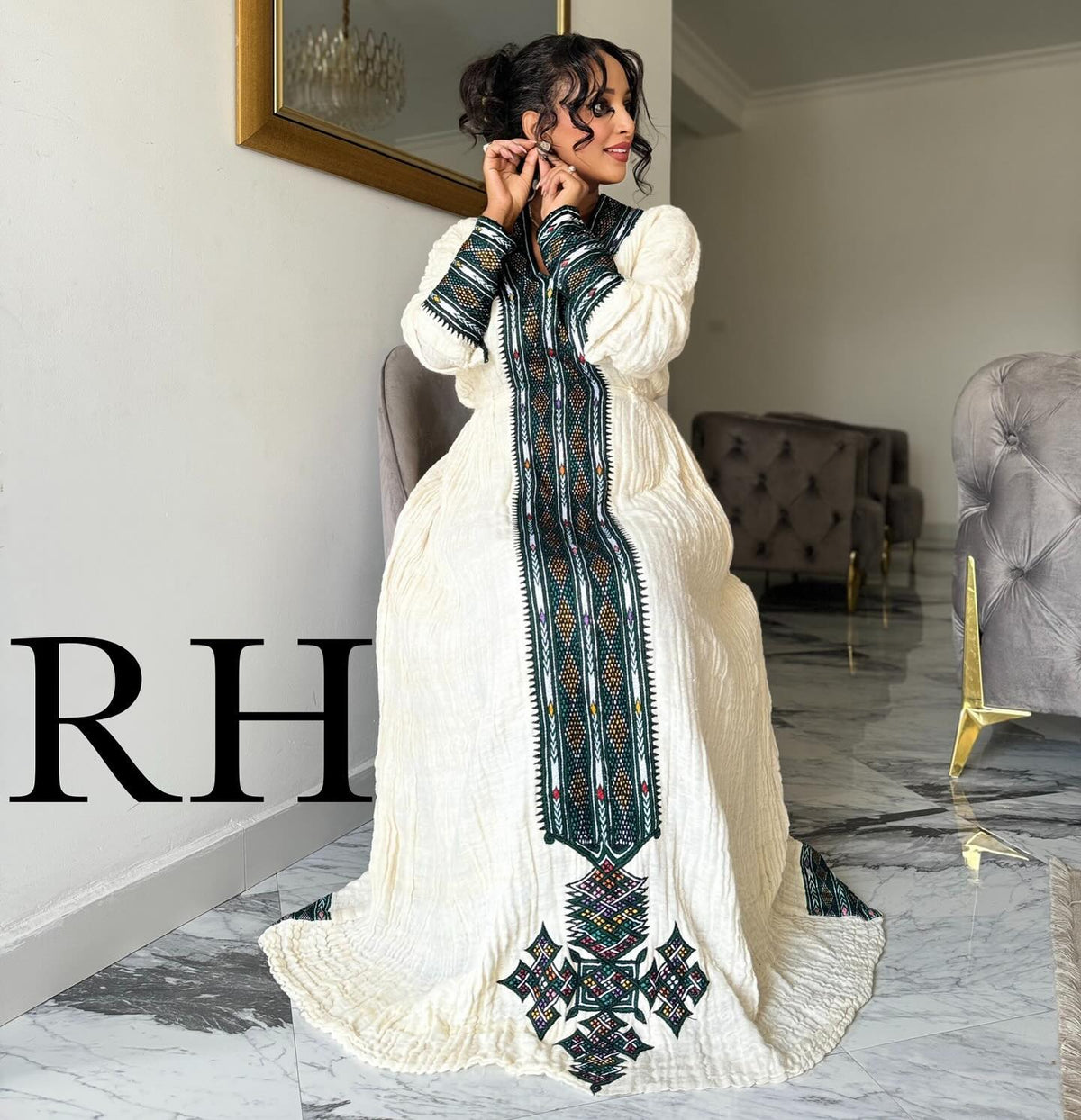 A woman models a long white embroidered Ethiopian National Dress in a modern living room setting. The dress features a high collar, long sleeves, and a prominent vertical panel with intricate green, black, and red geometric embroidery.