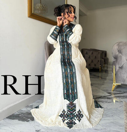 A woman models a long white embroidered Ethiopian National Dress in a modern living room setting. The dress features a high collar, long sleeves, and a prominent vertical panel with intricate green, black, and red geometric embroidery.