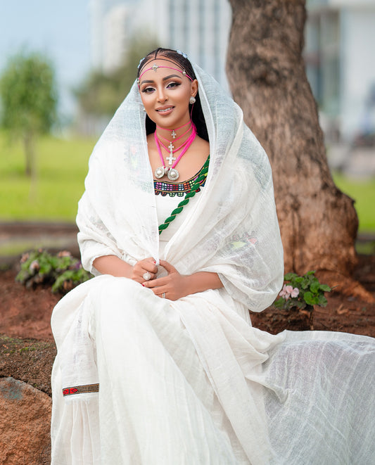 A woman wearing a traditional white Axum Fetil with a sheer white head covering and a green and red patterned neckline, seated outdoors near a tree trunk.