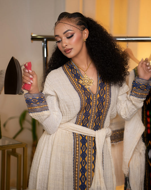 A woman modeling a cream-colored traditional Axum Fetil with blue and gold embroidered patterns, holding a pink iron in a boutique setting.