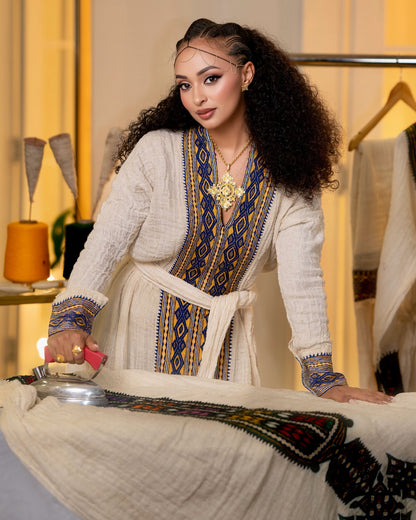 A woman with long curly dark hair is shown indoors, wearing a cream-colored traditional Axum Fetil with blue and gold embroidered patterns. She is ironing a matching cream-colored garment laid out on a table.
