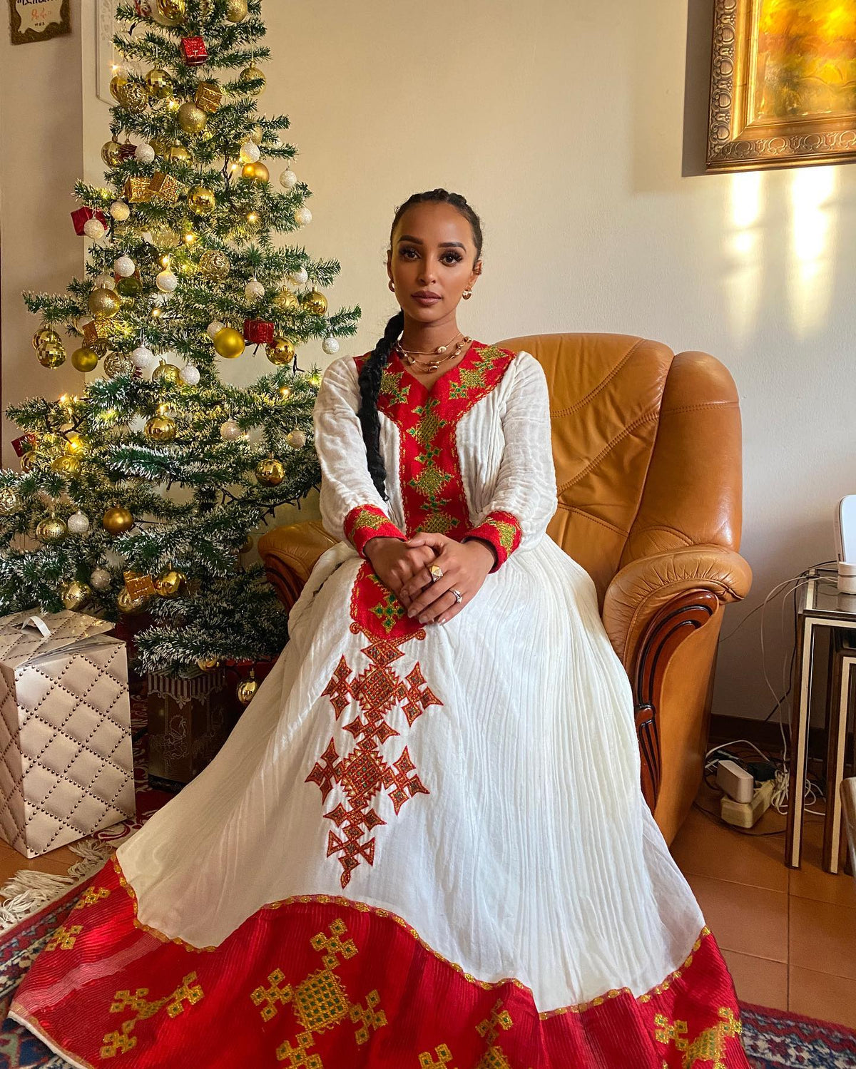 A woman wearing a traditional white and red embroidered Ethiopian National Dress sits in a brown leather armchair next to a decorated Christmas tree in a home setting.
