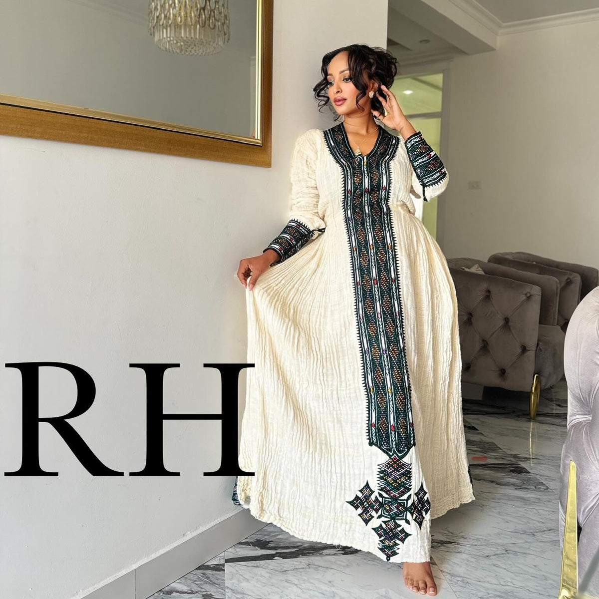 A woman models a long cream-colored Ethiopian National Dress with dark green and black geometric embroidery down the front and on the sleeves, standing in a modern home interior.