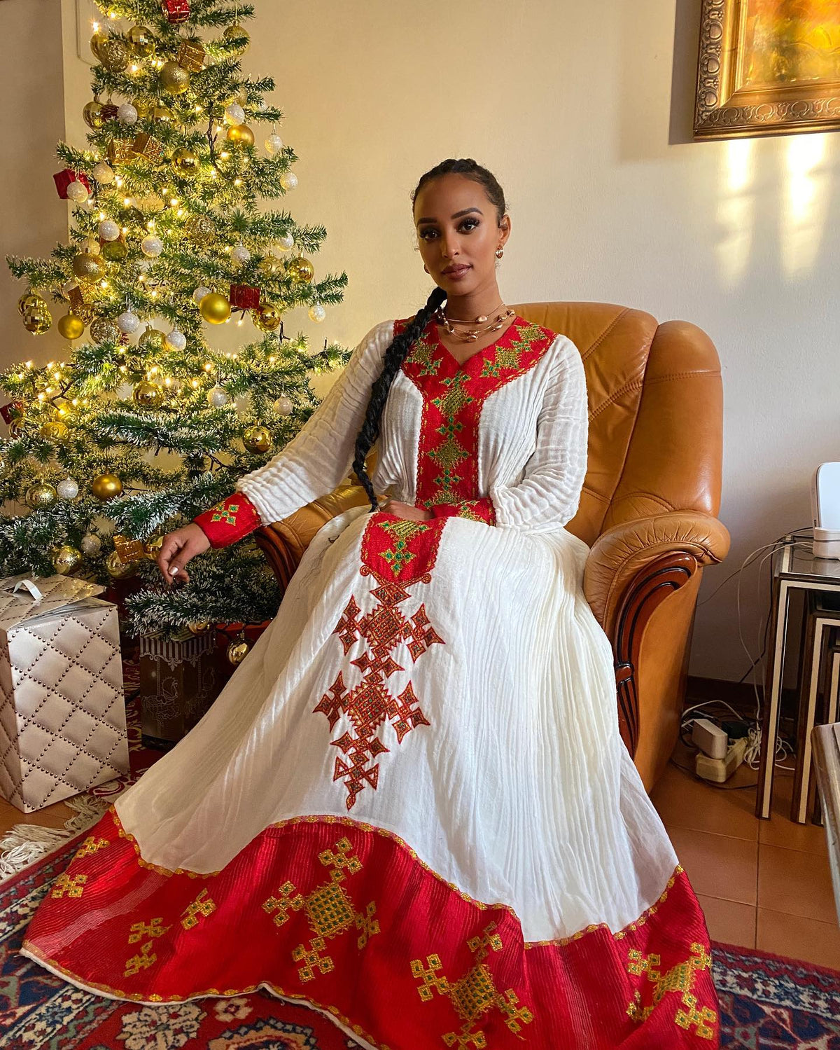 A woman wearing a traditional long white Ethiopian National Dress with red and green embroidered details sits in a brown leather armchair next to a decorated Christmas tree. The dress features a wide red hem with gold patterns.
