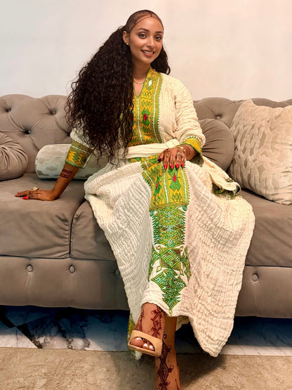 A woman with long curly black hair sits on a gray tufted sofa wearing a cream-colored traditional Axum Fetil with green and yellow embroidered details. She is wearing beige sandals and has henna on her legs.