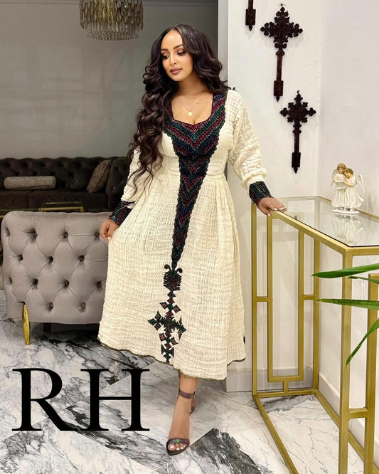 A woman models a long-sleeved cream-colored Ethiopian National Dress with a black and multicolor embroidered V-neckline and cross motifs down the front, standing in a modern living room setting.