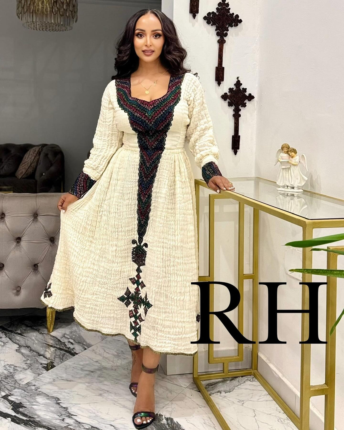 A woman models a long-sleeved cream-colored Ethiopian National Dress with intricate black, red, and green geometric embroidery concentrated on the V-neckline, front panel, and hem. She is standing in a modern interior setting with a gold console table and marble floor, with a large 'RH' watermark overlay on the bottom right.
