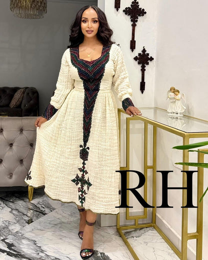 A woman models a long-sleeved cream-colored Ethiopian National Dress with intricate black, red, and green geometric embroidery concentrated on the V-neckline, front panel, and hem. She is standing in a modern interior setting with a gold console table and marble floor, with a large 'RH' watermark overlay on the bottom right.