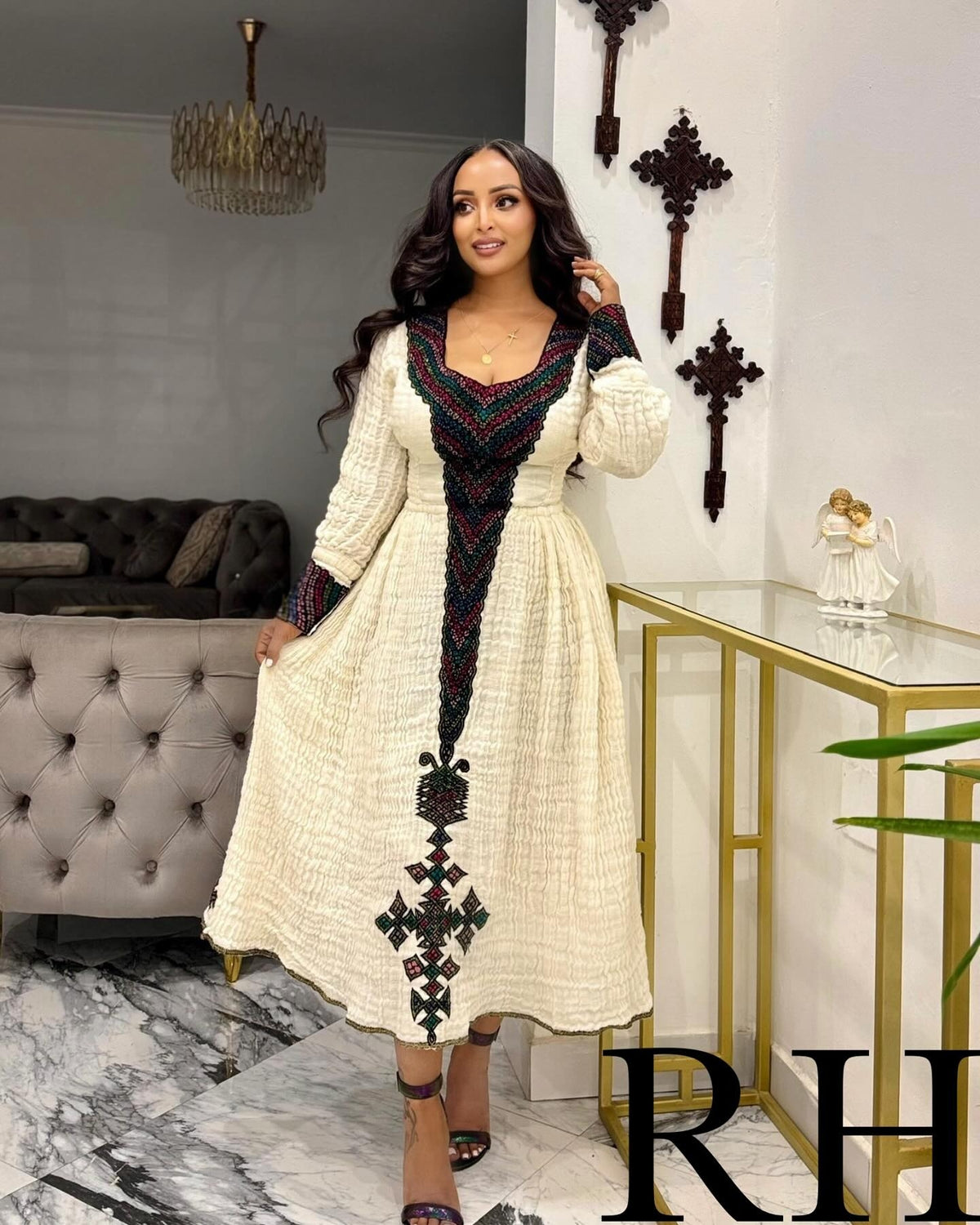A woman models a long-sleeved cream-colored Ethiopian National Dress with a black and red geometric embroidered pattern down the front and around the neckline, set in a modern living room environment.
