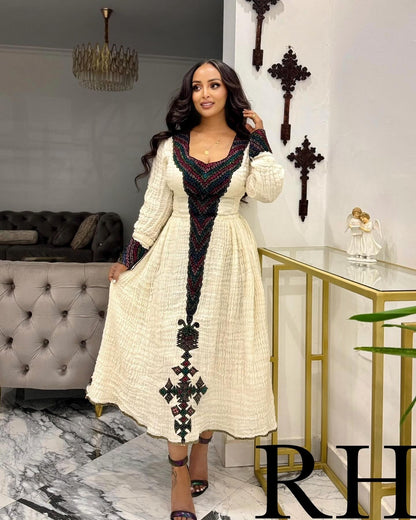 A woman models a long-sleeved cream-colored Ethiopian National Dress with a black and red geometric embroidered pattern down the front and around the neckline, set in a modern living room environment.