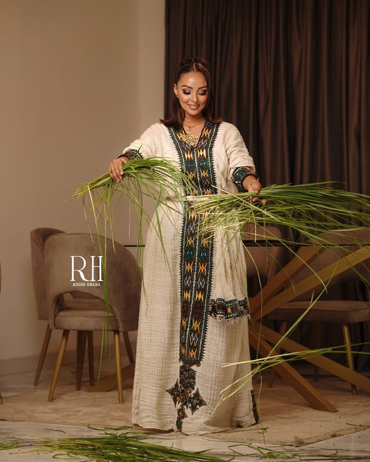 A woman modeling a long cream-colored embroidered Axum fetil while holding a bundle of green reeds in a styled indoor setting. The dress features intricate geometric embroidery in black, gold, and blue along the neckline, front panel, and hem.