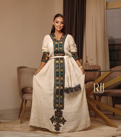 A woman modeling a long cream-colored Moroccan Axum fetil with colorful geometric embroidery in green, black, and gold. The dress features a V-neckline, long sleeves, a matching waist tie belt, and a draped dupatta.