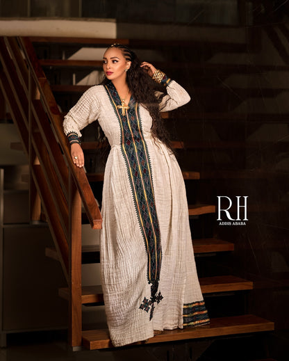 A woman wearing a long cream-colored textured Axum fetil with a dark blue, green, and gold embroidered vertical panel and matching embroidered cuffs, posing on wooden stairs. The brand logo 'RH AMIR KARIM' is visible on the right side.