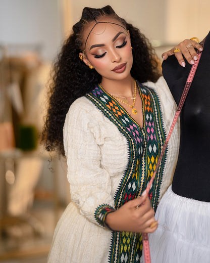 A woman with long curly hair models a cream-colored embroidered Ethiopian National Dress or tunic with a colorful geometric pattern along the neckline and sleeves, holding a pink measuring tape next to a black dress form.