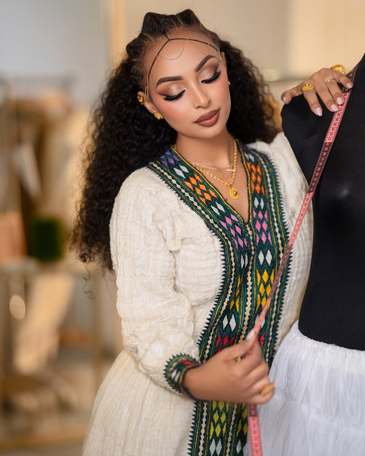 A woman with long curly hair models a cream-colored embroidered Ethiopian National Dress or tunic with a colorful geometric pattern along the neckline and sleeves, holding a pink measuring tape next to a black dress form.