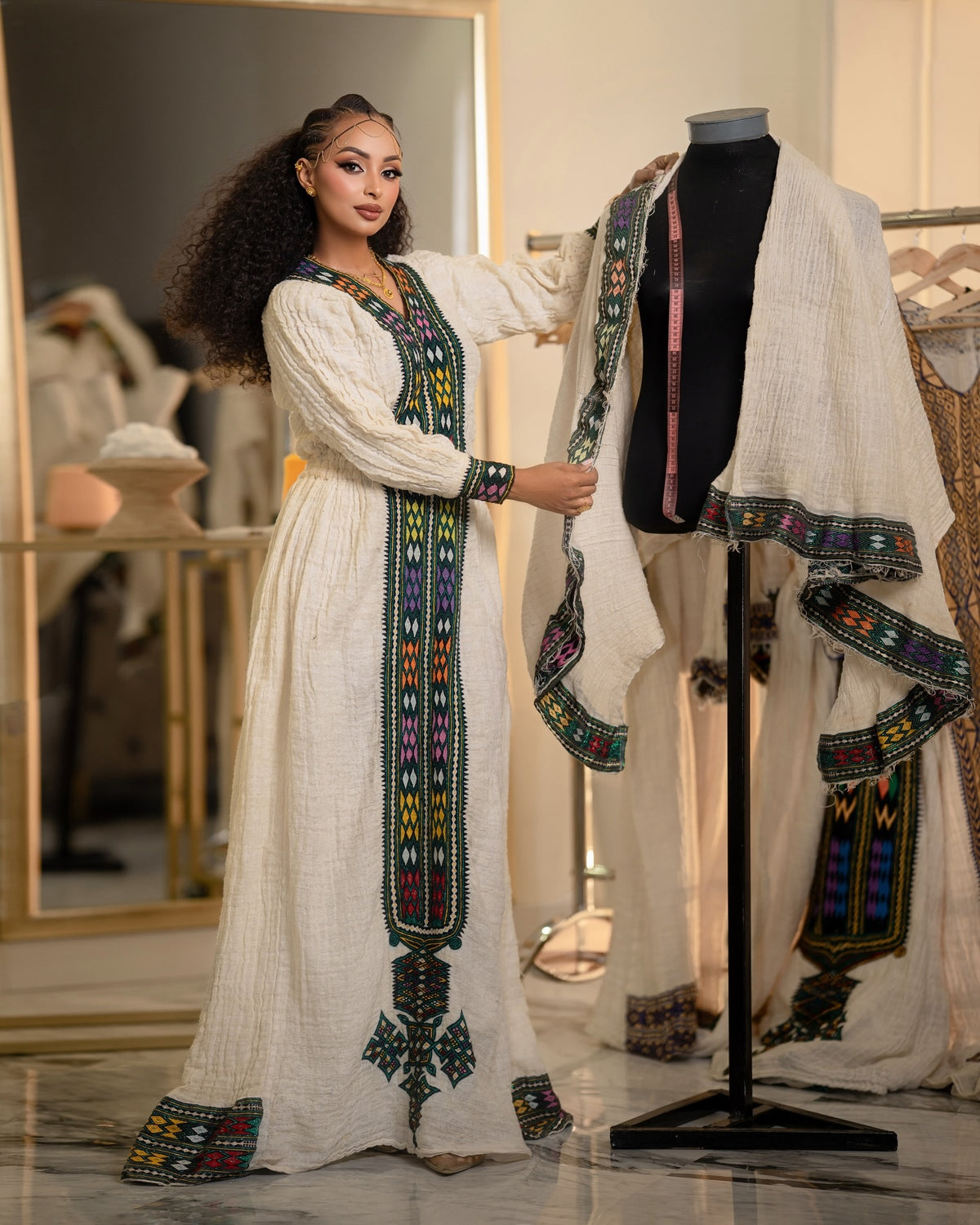 A woman stands in a boutique wearing a long white embroidered Ethiopian National Dress, while a mannequin displays a matching off-white embroidered garment draped over a black mannequin torso. The garments feature colorful geometric embroidery in green, blue, yellow, and red along the neckline, sleeves, and borders.