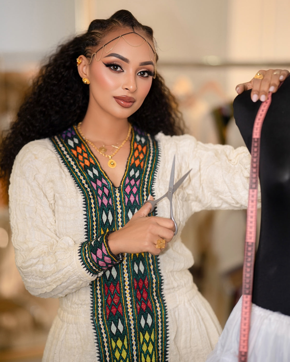 A woman with long curly hair is shown from the waist up, wearing a cream-colored embroidered Ethiopian National Dress with colorful geometric patterns down the front and on the sleeve. She is holding silver scissors and a pink measuring tape against a softly blurred indoor background.
