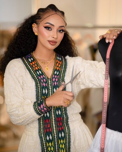 A woman with long curly hair is shown from the waist up, wearing a cream-colored embroidered Ethiopian National Dress with colorful geometric patterns down the front and on the sleeve. She is holding silver scissors and a pink measuring tape against a softly blurred indoor background.