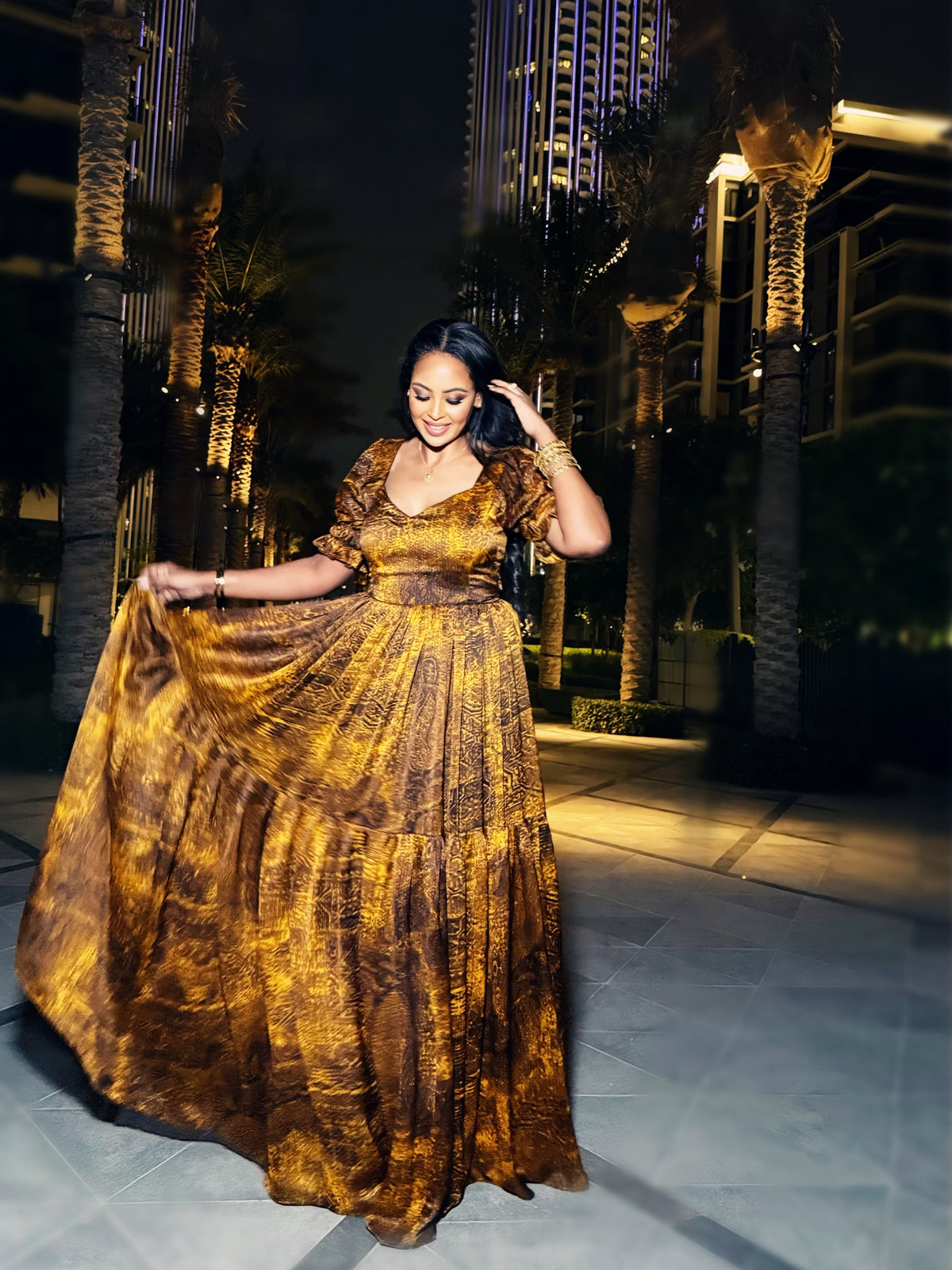 A woman wearing a voluminous, floor-length Habesha Dress  with a textured metallic gold and bronze pattern, holding the skirt outwards against a dark outdoor background at night.