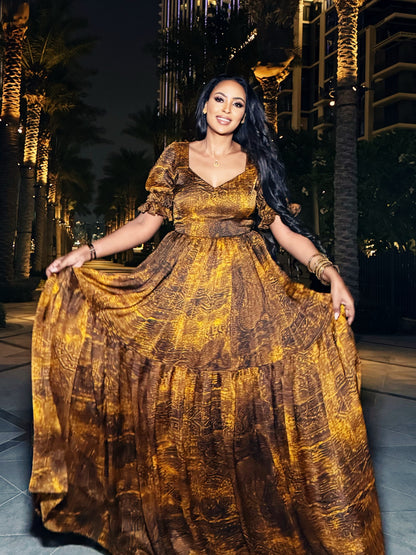 A woman wearing a voluminous gold and brown patterned maxi Habesha Dress  with short puff sleeves, holding out the skirt to show its full tiered flow, set against a nighttime urban background with illuminated palm trees and buildings.