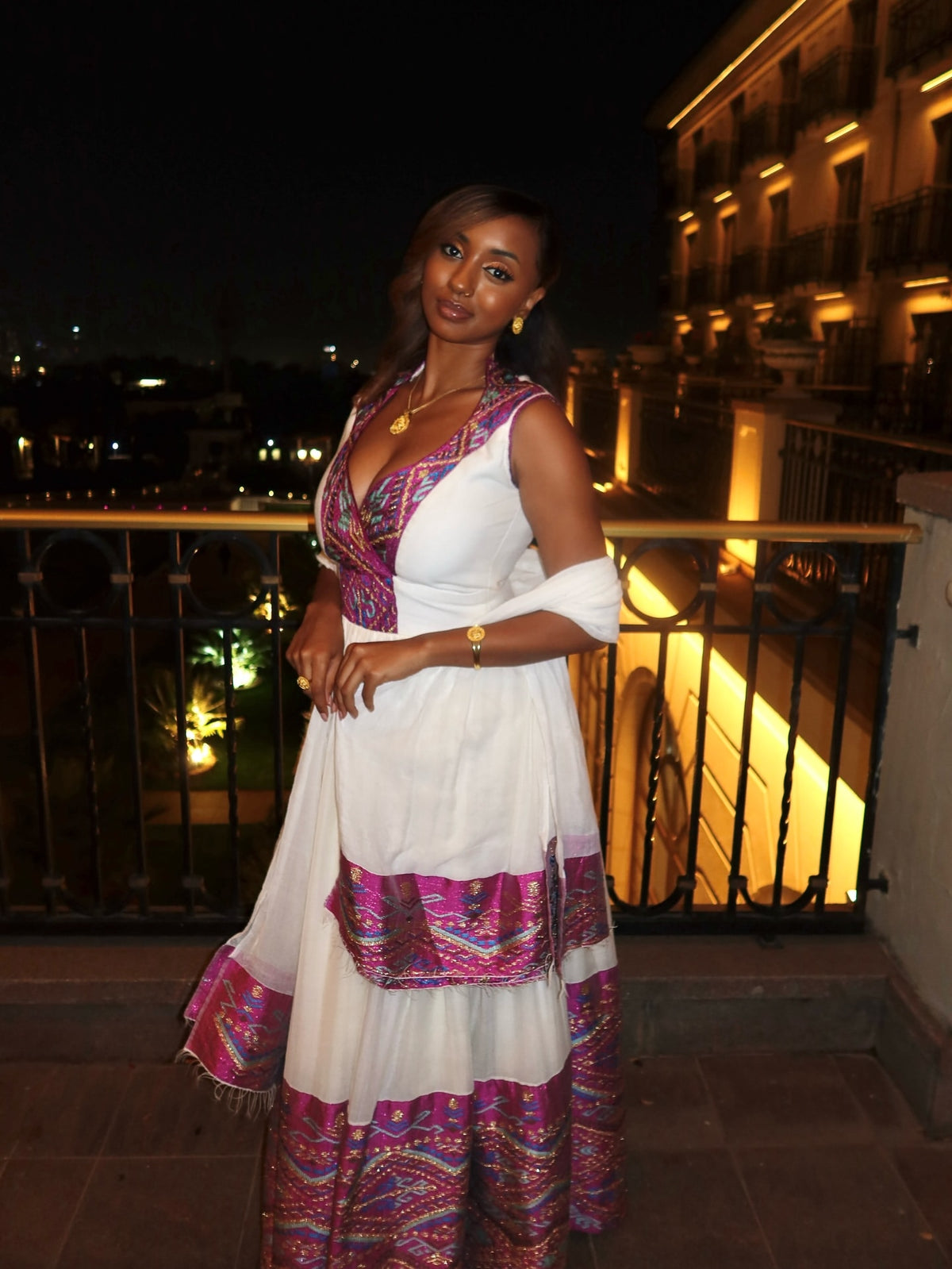Woman in a colorful Habesha Wedding Dress standing on a balcony at night.
