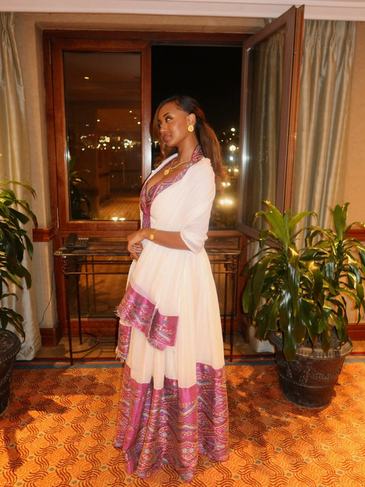 A woman wearing a white maxi Habesha Wedding Dress with a pink and purple patterned border and neckline, standing in an indoor setting with a window and potted plants.