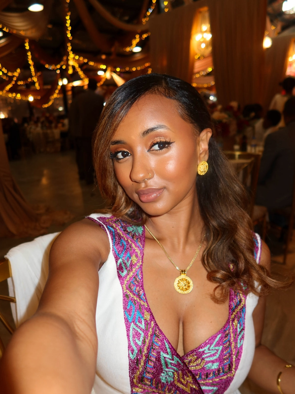 A woman taking a selfie in a decorated event hall, wearing a white sleeveless Habesha Wedding Dress with a vibrant purple, pink, and blue patterned V-neckline and gold jewelry.