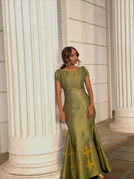 A woman wearing a floor-length olive green Habesha zuria  with short puff sleeves and yellow embroidered details near the hem, standing between large white classical columns.