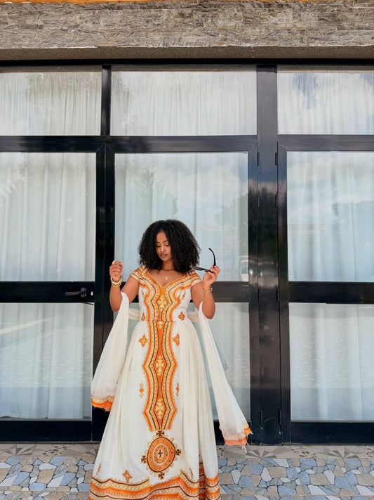 A woman stands outdoors in front of a building with large black-framed glass doors and white curtains, wearing a long white traditional Ethiopian national dress with prominent orange and gold embroidery and a matching sheer white shawl.