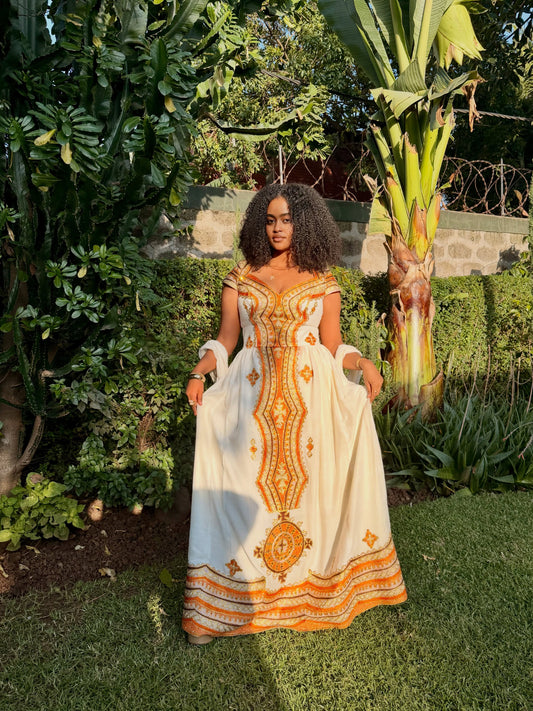 A woman stands outdoors in a lush garden wearing a long white Ethiopian national dress with prominent orange and yellow embroidered patterns. The dress features a V-neckline, cap sleeves, and a full skirt with decorative geometric and floral embroidery concentrated on the front panel and hem.