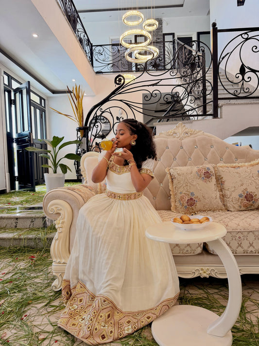 A woman in a white and gold traditional Ethiopian Shop Near Me sits on an ornate cream sofa in a luxurious interior, holding a glass of orange juice next to a small white side table.