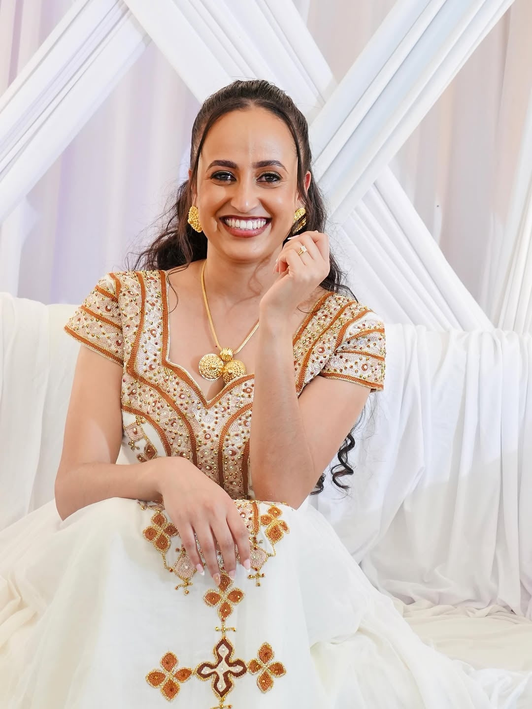 Woman wearing a white and gold embroidered traditional Habesha Kemis with a decorative background.